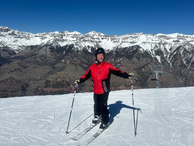Ken Corriveau on skis with arms wide on a mountain summit